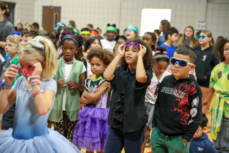 Children in costumes at a school event, some looking up towards camera, others standing around.