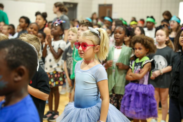 Little girl in blue dress standing in front of group of children wearing costumes.