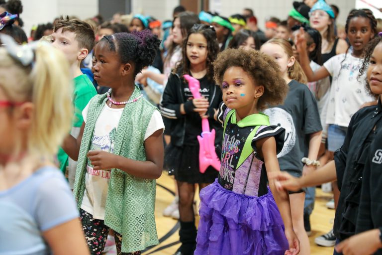 Children in costumes at a school event, some holding signs, all smiling and engaged.