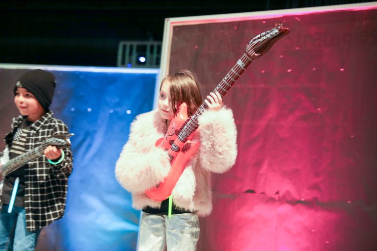 Two children playing guitars on stage, one in front of a backdrop with a starry sky and the other in front of a red curtain.