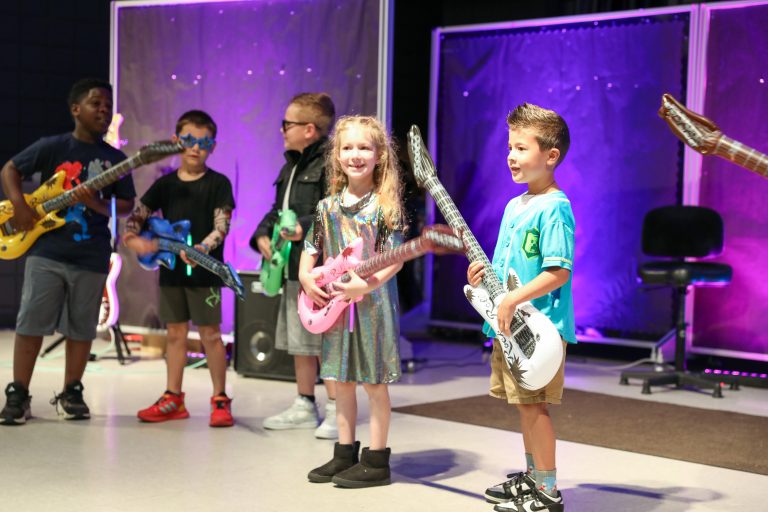 Children on stage holding guitars, smiling at camera.