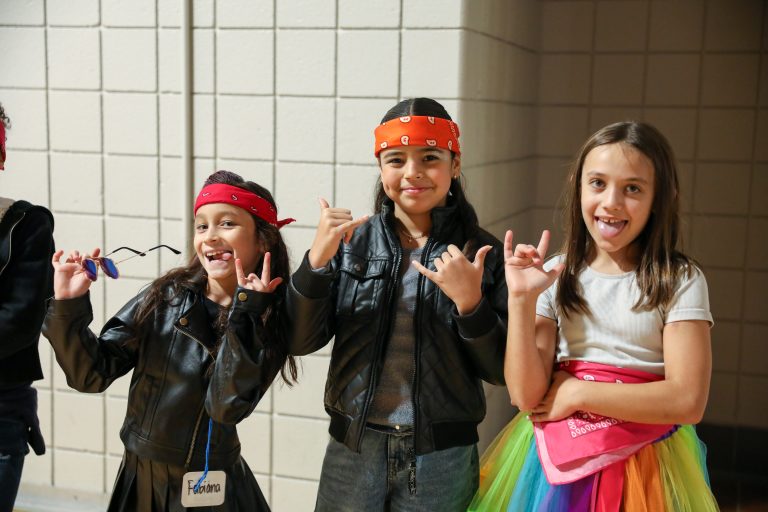 Three children posing in front of a building, one making a peace sign, another wearing sunglasses, and a third holding a