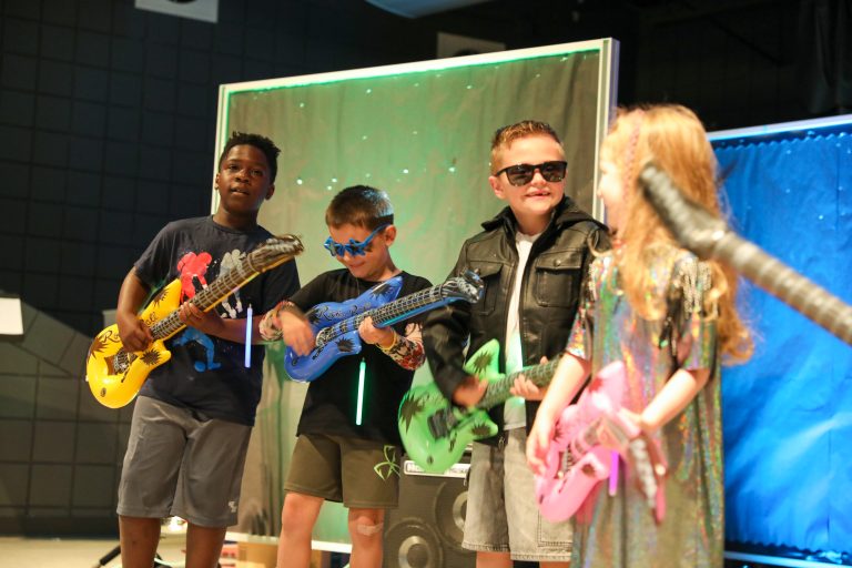 Three children posing with a guitarist in front of a stage backdrop.