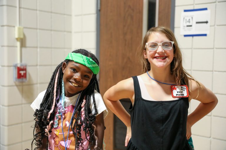Two young girls posing in a hallway, one wearing a blue headband and glasses, the other with braided hair and a bandana,