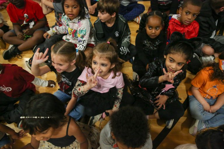 Group of children sitting on gym floor, some holding hands, all looking at camera, smiling and posing for picture.