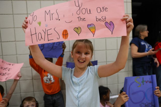 Girl holding sign that says "you're the best Mrs Hayes" in a gymnasium.