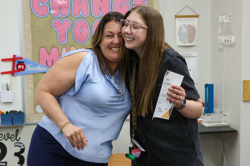 Two people standing close together, smiling at the camera, one person holding a piece of paper. They are in an indoor