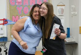 Two people standing close together, smiling at the camera, one person holding a piece of paper. They are in an indoor