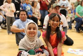 Two young girls in traditional Indian attire sitting on floor at event, smiling towards camera.
