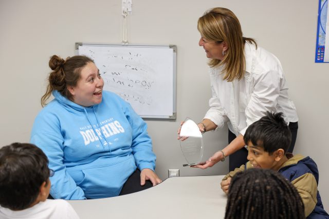 Woman presenting to children in a classroom setting, holding up a small object for demonstration.