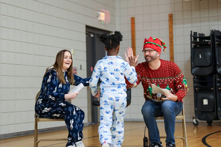 Two people sitting on a chair in front of a gymnasium, one waving at the camera and the other holding a child's hand.