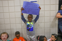 A young boy holding a sign in front of him, standing next to other children in a room. The sign has writing on it.